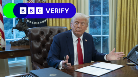 Donald Trump sitting at his desk in the Oval Office holding a pen. He is wearing a dark blue suit with a US flag badge on it, a white shirt and a red tie. The BBC Verify logo has been added to the top left corner of the image.