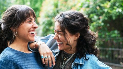 Two friends laugh together with a park in the background.