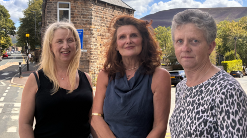 Three women stand together and smile while standing by a roadside on a sunny day. Clouds can be seen in the sky with trees and the view of a rural headland seen in the distance