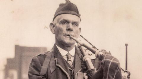 A sepia-hued image of piper Daniel Laidlaw, a smart older man in uniform, playing the bagpipes. He is white-haired, with a Glengarry hat and a white winged moustache.