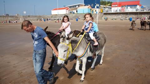 A man leads a pair of donkeys along a sandy beach as two kids sit on each of them. They are people sat on the beach with colourful buildings in the background.