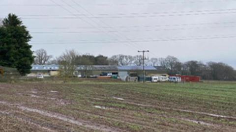 Farm land with long rural buildings in the distance and several caravans. 