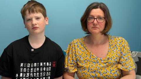 Shows a woman and young boy looking into the camera with a serious expression. The woman has short brown hair and is wearing glasses and a yellow and blue floral shirt. The boy has red hair and is wearing a black patterned shirt.