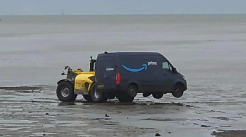 A forklift truck, or similar vehicle, is lifting a navy blue Amazon Prime branded van on mudflats. The sea is behind, and there is blue sky