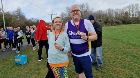 Joe Baker is standing on the right, on green grass. He has white hair and black glasses. He is wearing a white top with a purple vest that says "Stroke Association". He has a medal around his neck. To his right is a woman with a blonde bob. She is wearing a blue top and rainbow cheetah print leggings, and also has a medal around her neck