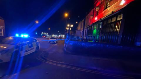 A street with a police car parked outside a pub, on the corner of a road - with a cordon in place