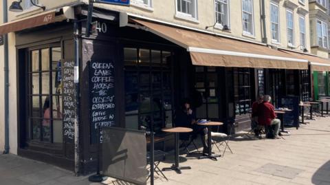 The sun is shining on tables and chairs outside a cafe. A faded brown canopy hangs over the front of the cafe. 
White chalk writing on a black sign outside the coffee house reads Queen's Lane Coffee House "the oldest established coffee house in Europe since 1654. An older couple sit at one of the tables at the far corner of the image