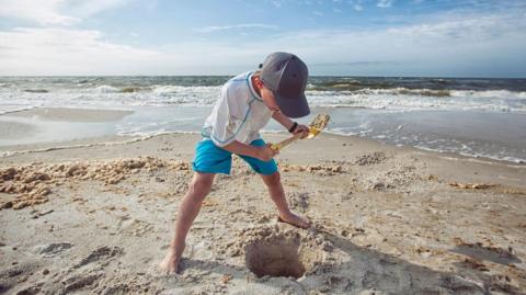 A young boy digging a hole in the sand on a beach. He has a yellow plastic spade and a is wearing a blue baseball-style cap.