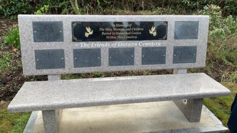 A stone bench is set on a concrete slab. It has a number of plaques on it. The one at the top reads: 'In memory of the men, women and children buried in unmarked graves within this cemetery'