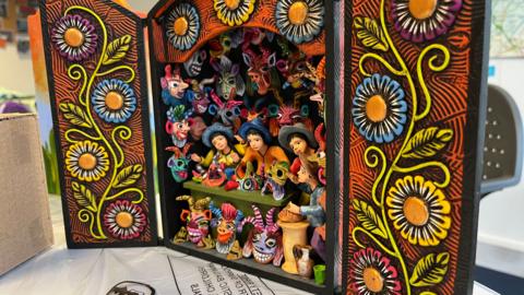 A  Peruvian Retablo that has been created at the March workshop. Its is beautifully painted and looks almost like a small cabinet with the doors open. On either side of the door is painted yellow, blue and pink flowers. Inside is models of three people making masks. They are surrounded by other model masks making a very bright and vivid scene. 