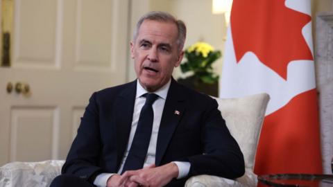 Man sits with his hands interlaced and Canadian flag in the background