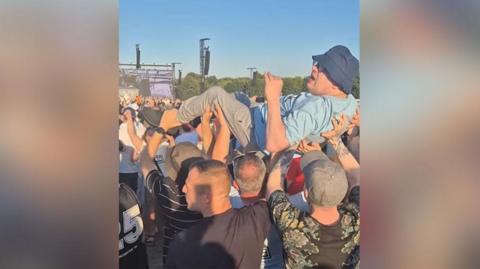 Daniel Tester, wearing a sky blue t-shirt and navy blue bucket hat, is held up in the air at an Oasis gig at Heaton Park by fellow fans.