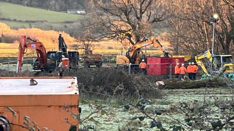 Workers operate diggers and move crates in a field to try and repair a burst water pipe.