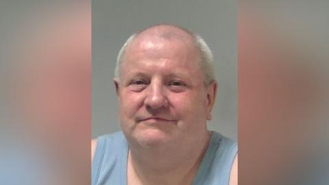 A custody photo of Steve Wright who is smiling at the camera. He is wearing a light blue vest and is bald with some grey hair around the side of his head.
