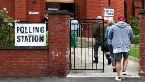 People arrive to vote at a polling station in the Makerfield by-election. Polling station signs are on the walls of a red brick building