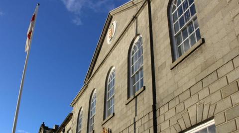 A Guernsey flag and blue sky stands next to a building with grey bricks and blue windows, showing the States logo. 