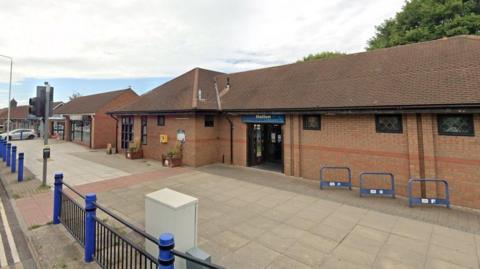 A single-storey building in red brick with a low sloping roof. There is a paved area in front of the building with blue bicycle racks and a blue and black fence.