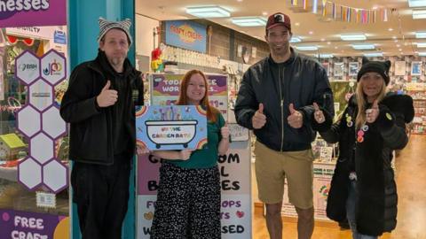 Dave, a man wearing all black clothes and a hat, standing next to a woman who works in the shop - wearing a green top and a printed skirt. David James is wearing tan-coloured trousers and a black zipped jacket, Kez Hayes-Palmer wearing a black coat. They are all standing outside the Unity store showing a thumbs up to the camera. 
