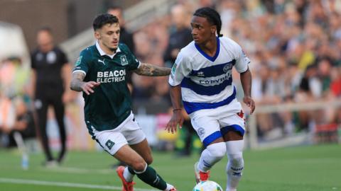 Putman running with the ball, looking to his right, wearing QPR home blue and white hoops, holding off an attacking Plymouth Argyle player in green.