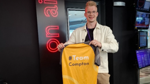 Tom is stood outside Radio Manchester studios holding a gold football shirt with Team Compton on the back. He has a cream jacket on over a dark top and brown trousers.