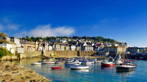 sailing boats in a harbour surrounded by houses, under a blue sky