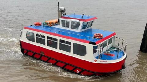 The Knott End Ferry crossing the brown waters of the River Wyre. It is a relatively small boat with a red bottom, white frames windows and a blue deck.