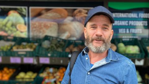 A man with a blue shirt and hat speaking to the camera with an array of fruit and veg behind him in a soft focus