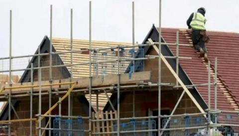 Stock image of a house being built, with scaffolding and a builder with a high vis jacket on the roof doing work