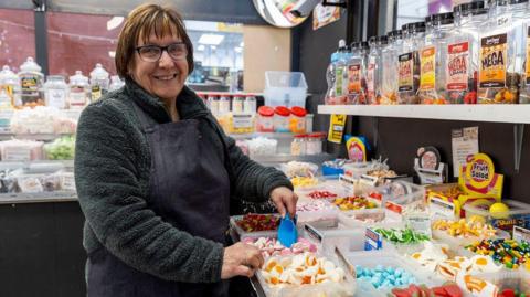 A woman with a brown bob and glasses is wearing a dark green jumper and a black apron. She is standing in a sweet shop, surrounded by tubs of various sweets. She is holding a blue scoop which she is putting inside a box of pink and white mushroom-shaped sweets