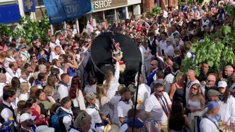 A crowd of people cheer and sing as a large black Obby Oss with blue ribbons dances through the streets of Padstow.