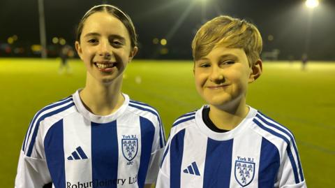 Two young girls wearing blue and white vertically striped football shirts are smiling at the camera.
There is a green football pitch behind them with floodlights.