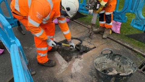 Two workmen work on a pothole. One is scooping out broken up tarmac with a shovel, while another works with a pneumatic drill to break up the remaining tarmac