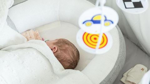 A baby sleeping in a cot with a mobile above. The baby is covered by a white blanket, so you can just see its head. The mobile features a colourful red and yellow swirl and a blue car