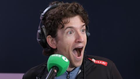 Greg James with his mouth open in disbelief in front of a radio microphone
