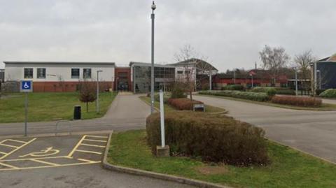 A low-lying school building with a two-storey glazed front and a road leading to the front.