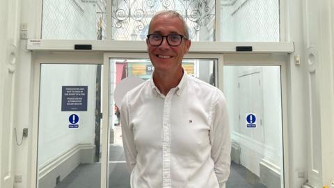 Simon Youden is standing in front of a glass door entrance. He is wearing a white button up shirt. He is holding his arms behind his back.