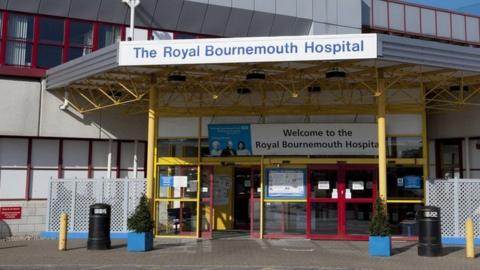 The front entrance of The Royal Bournemouth Hospital. The red, yellow and blue framed structure has automatic doors beneath signs for the hospital.