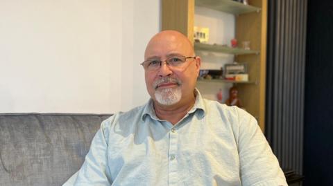 A man with a shaved head, glasses and a grey goatee-style beard. He is wearing a light linen shirt with the top-button unbuttoned. He is sitting on a dark grey sofa, in front of a wooden shelving unit. He is staring directly at the camera.