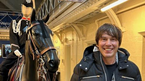 A smiling Professor Brian Cox standing in front of a model of a black horse with a mounted police officer mannequin on it. He is smiling and wearing a black padded coat. The background shows prison cells lit with yellow light