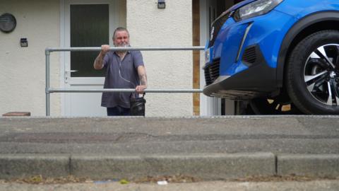 Michael Partridge, wearing a blue shirt and black trousers, stands outside his house in Medway, leaning on a metal barrier and behind a steep kerb. His white front door is behind him and his blue car is parked on his left.