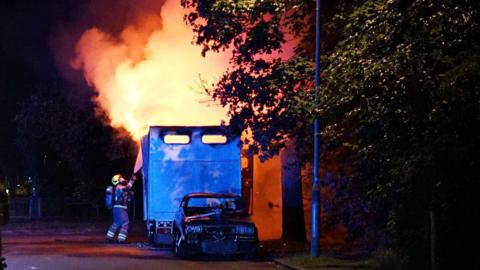 A firefighter uses a hose to try and get flames coming from a small white van under control.