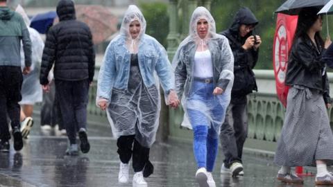 Two women in plastic overalls walk in the rain across a bridge in London.