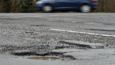 A pothole is at the bottom left of the image and has a small amount of water in it. There is a blue car in the background which is driving on part of the road.