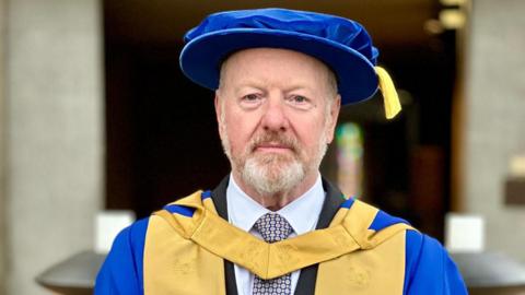 Sir Alan Bates wearing a blue hat with gold and blue robes. 