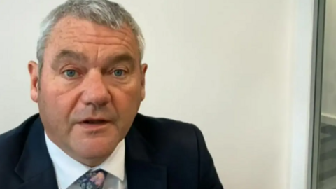 Mr Walker is wearing a dark suit, white shirt and pink tie. He has short grey hair.