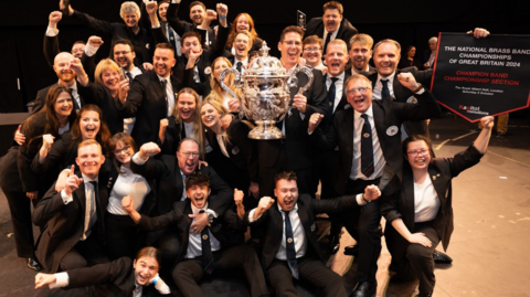 The band all looking ecstatic with their hands in the air and smiling with the conductor in the centre holding an enormous silver trophy