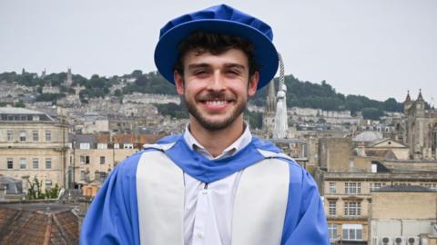 George Clarke is standing on a balcony overlooking the city of Bath. He is dressed in a graduation cap and gown after being given an Honorary Doctorate of Arts. The gown and cap are blue and silver. George has short dark hair and facial hair and is standing with his hands clasped in front of him, looking at the camera and smiling.