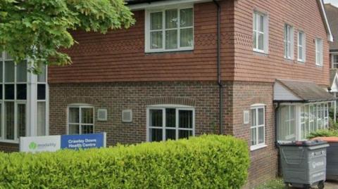 A tile and red brick building with white window frames behind a green hedge. A sign reads "Modality: Crawley Down Health Centre" 