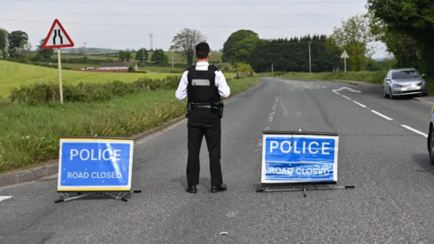 Police officer facing away from camera standing in the middle of the road with two signs that read "Police Road Closed"