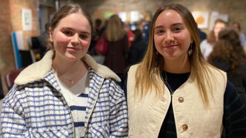 Katrina is standing on the left of the image. She is wearing a light blue and white chequed jacket. She has dark brown hair which is tied back in a ponytail. Amelie is standing on the right of the image. She is wearing a cream gilet jacket. She has light brown medium length hair.